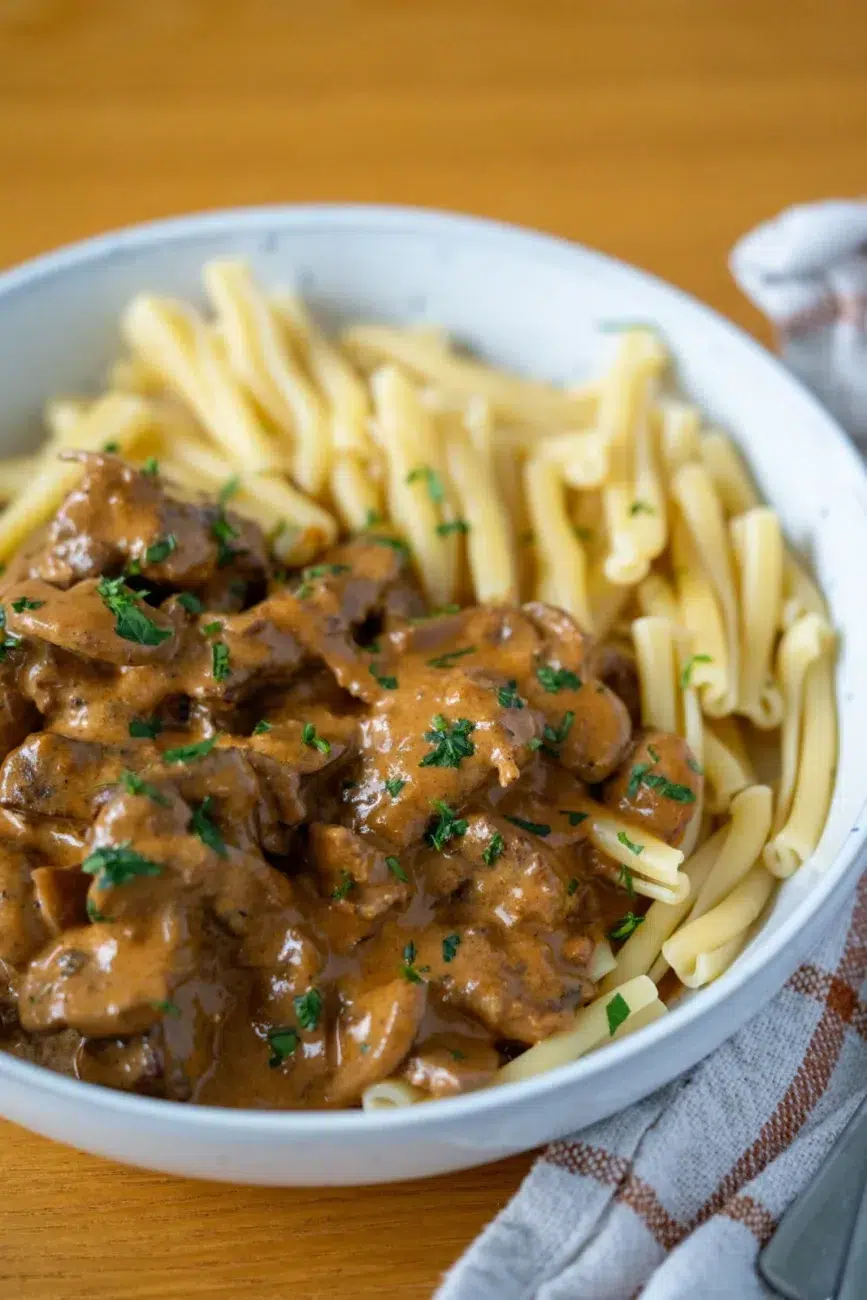 Angled close-up of a white bowl filled with creamy beef stroganoff and pasta, featuring tender beef pieces in a rich mushroom sauce, garnished with chopped parsley and paired with twisted pasta noodles, served on a wooden table with a checkered kitchen towel and cutlery on the side.