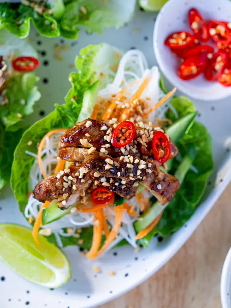 Close-up top view of a hoisin chicken lettuce cup filled with vermicelli noodles, julienned carrots, cucumber, crushed peanuts, and sliced red chili, served with a lime wedge and a bowl of fresh chili on the side.