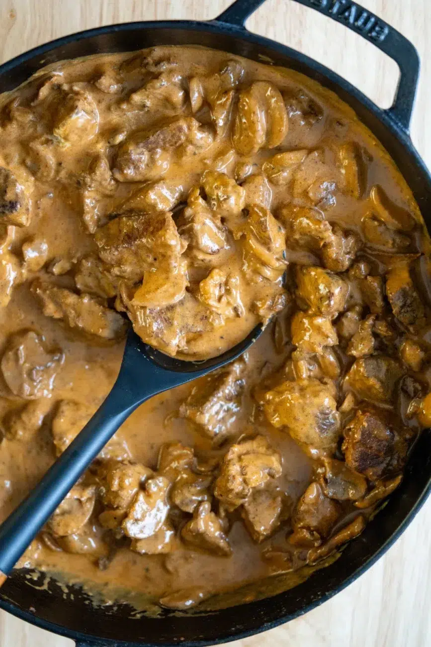 Overhead view of a cast iron pan filled with creamy slow-cooked beef stroganoff, featuring tender chunks of beef and sliced mushrooms coated in a rich, velvety brown sauce, with a black serving spoon lifting a portion from the pan.