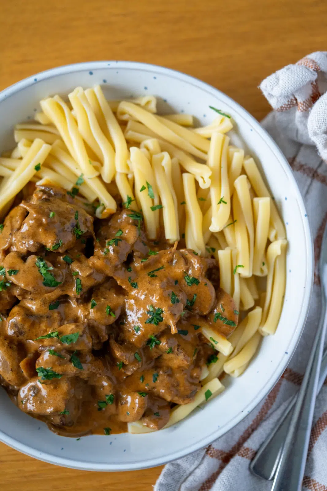 Top view of a white speckled bowl filled with slow-cooked beef stroganoff and pasta, showcasing tender beef in a rich creamy sauce alongside twisted pasta noodles, garnished with chopped parsley and served on a wooden table with a checkered napkin and silver cutlery.