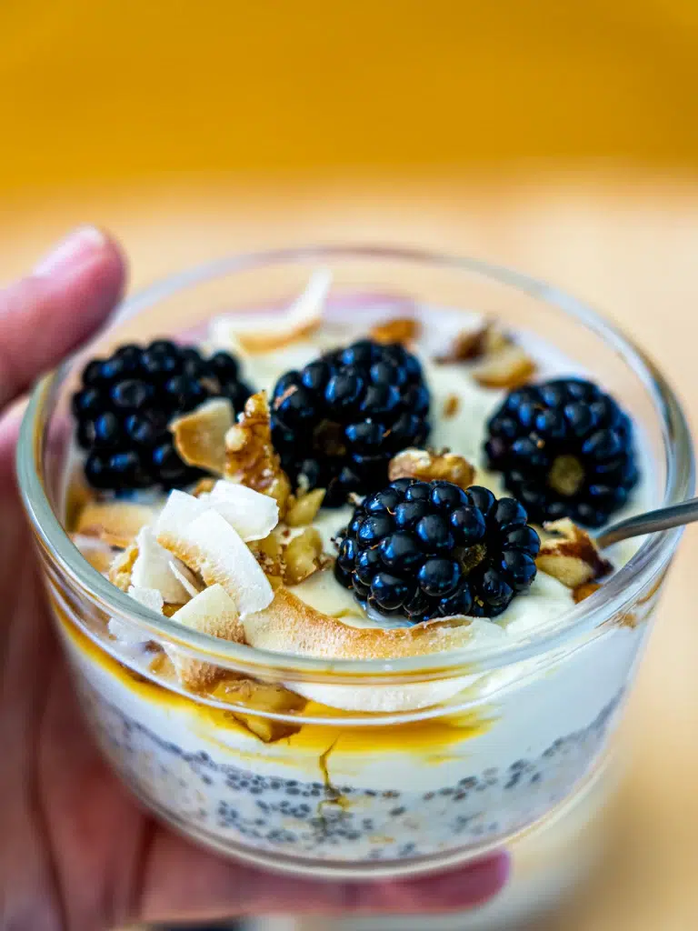 Holding a glass of creamy chia and oat pudding, finished with fresh blueberries and shredded coconut.