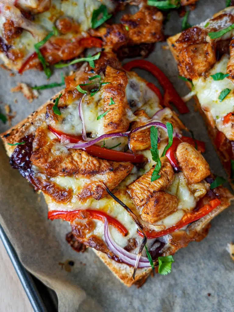 Close-up of a cheesy chicken flatbreads topped with grilled chicken pieces, red bell peppers, red onions, and fresh parsley on a baking tray