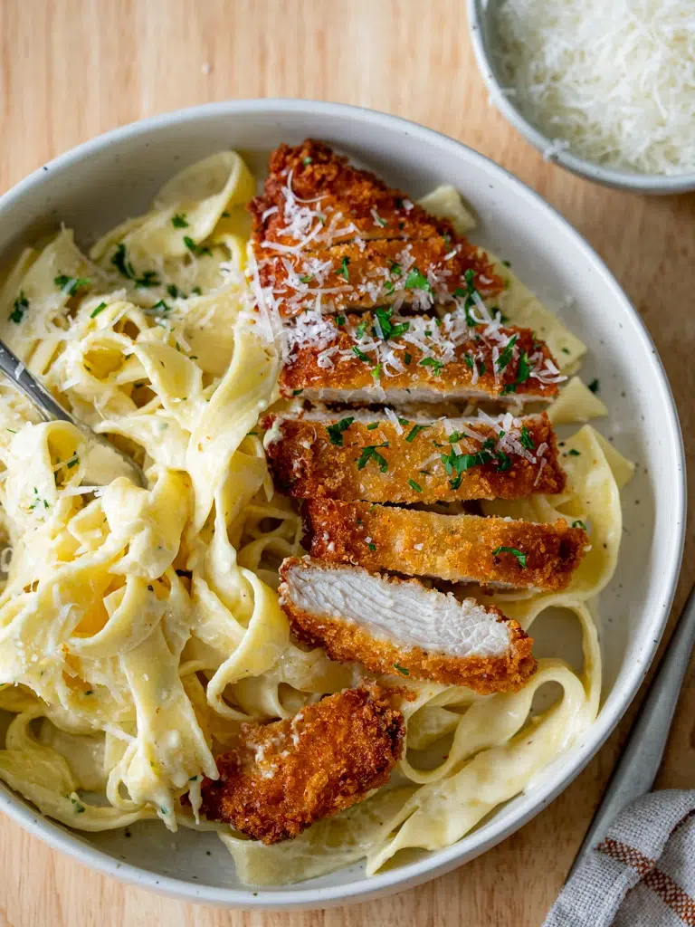 Top view of sliced crispy chicken on fettuccine Alfredo with a fork resting beside the plate