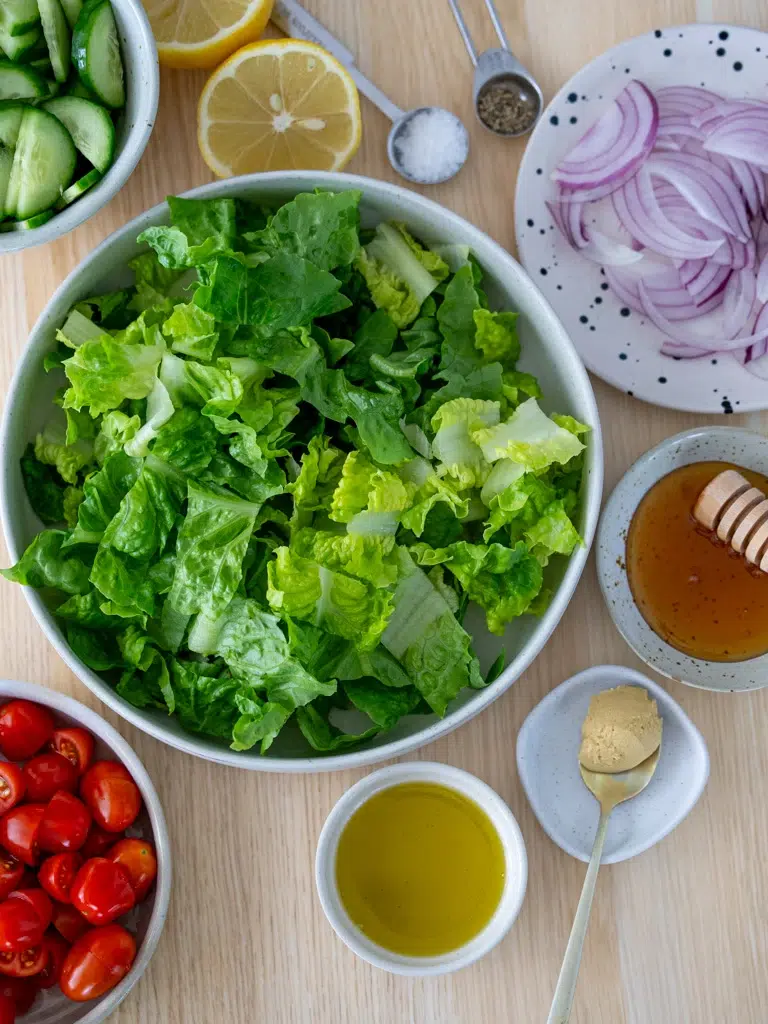 Flat lay of fresh salad ingredients including romaine lettuce, cherry tomatoes, cucumber, red onion, lemon, olive oil, honey, mustard, salt, and pepper on a wooden surface.
