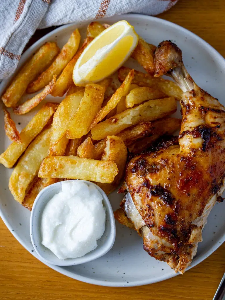 Crispy oven fries with grilled chicken leg, lemon wedge, and garlic sauce on a white plate.