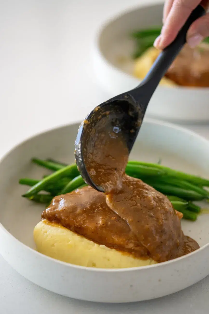 Gravy being spooned over pork chop on a bed of mashed potatoes, served with green beans in a shallow white bowl