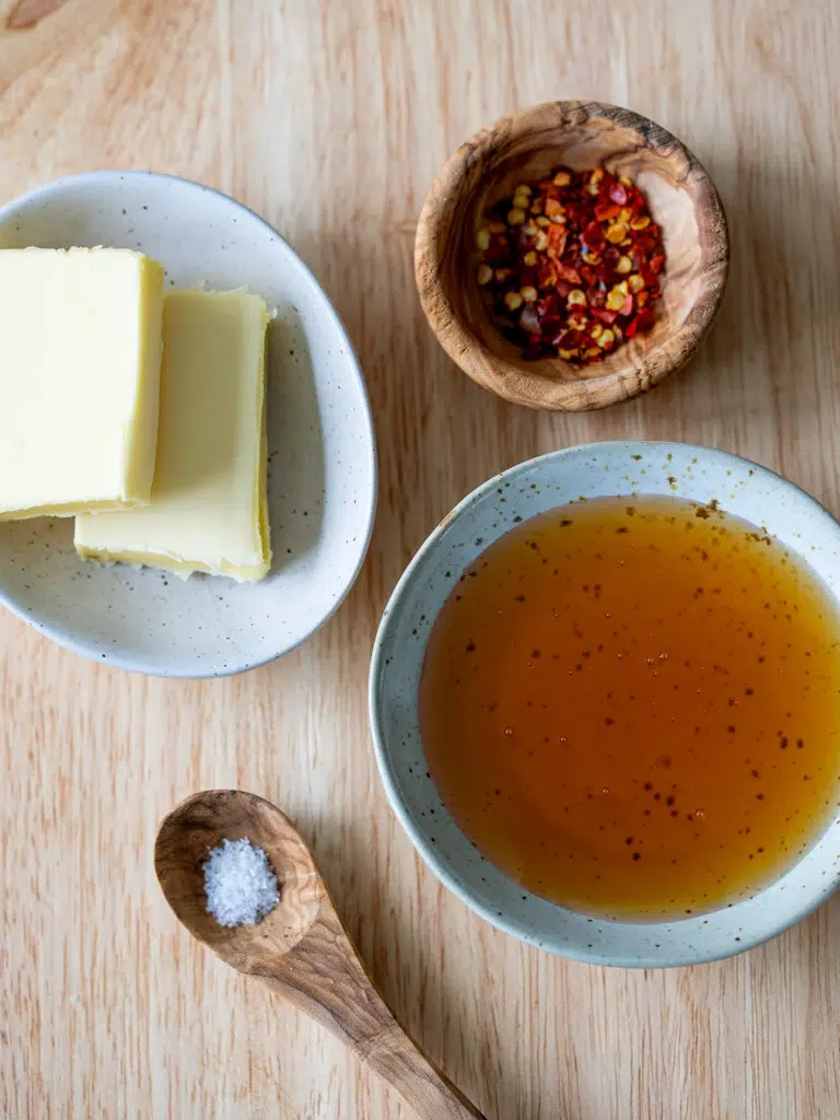 Ingredients for easy hot honey sauce on a wooden surface, including butter, chili flakes, honey, and salt displayed in small bowls and a wooden spoon.
