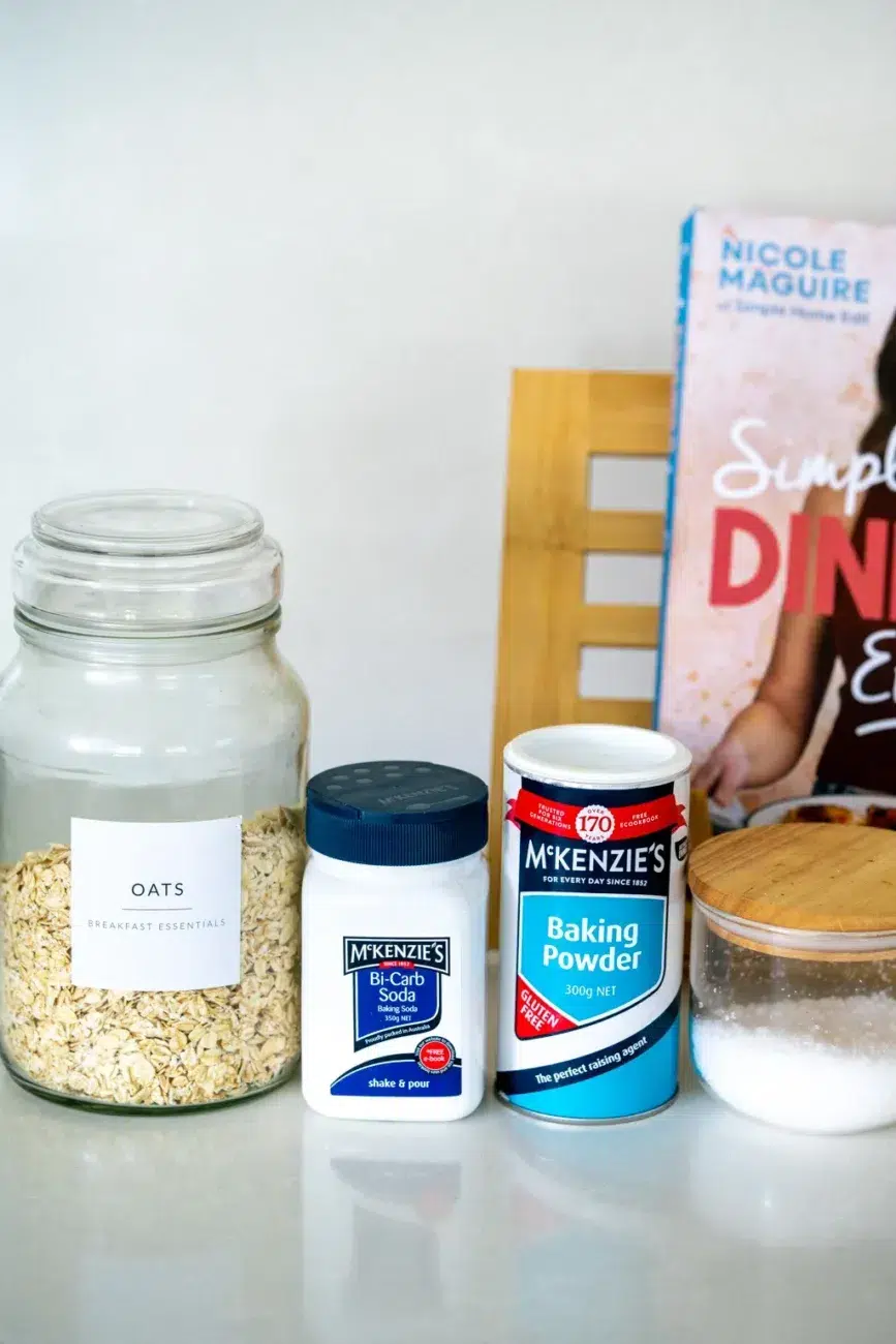 Dry ingredients for lunchbox protein pancakes displayed on a kitchen counter, including a large jar of oats labeled 'Breakfast Essentials', containers of McKenzie's bi-carb soda and baking powder, a small jar of sugar with a wooden lid, and a cookbook titled 'Simple Dinner Edit' by Nicole Maguire in the background.